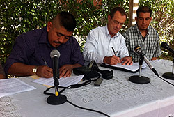 Three men sitting at a table with microphones, while two write on paper.