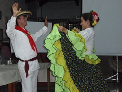 A  man in white garmets with a red neck sash and a woman in green, yellow  and polka dotted attire perform traditional dances.