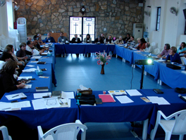 A group of people seated at tables with blue tablecloths arranged in a rectangle in a room.