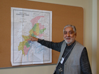 A man wearing a string name badge in grey vest and black shirt point to a map of Pakistan on a wall.