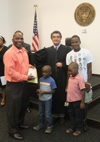 A man stands beside his children and a judge in his cloak, holding a citizenship document. An American flag and seal is behind them.