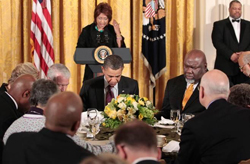 A woman delivers an invocation from a podium, while Barack Obama and other religious figures are seated in front of it, praying.