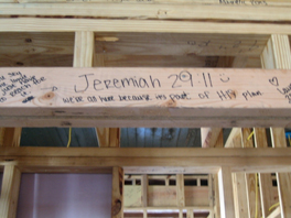 A wooden sign with a graffiti dedication hanging from the wooden frame of a house.