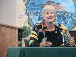 The Rev. Jane Adams Spahr, sitting on the stand during trial in front of a mural painted with a ship sailing on water, speaking.