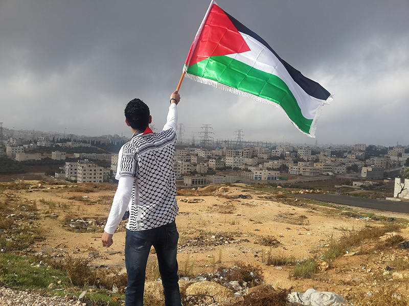Image of man waving Palestinian Flag over Palestine. Photo by Ahmed Abu Hameeda - Unsplash.