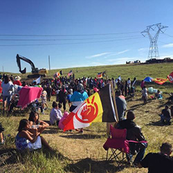 Protesters congregate next to a construction site for the Dakota Access Pipeline on Monday morning, as a crew arrives with machinery and materials to begin cutting a work road into the hillside. The flag in the foreground belongs to the American Indian Movement. (Photo by Daniella Zalcman)