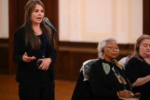 A woman stands and speaks at a microphone while others listen.