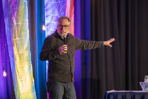 A man speaks with a handheld microphone on a stage with a colorful backdrop.
