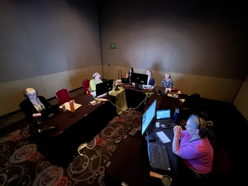 A group of people sit in a large dark room, their faces illuminated by computer screens. 