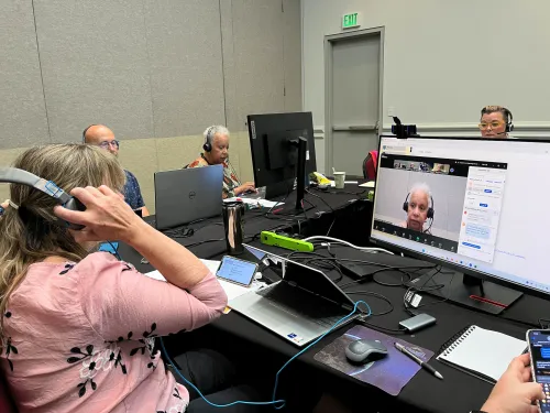 A woman with headphones looks at a desktop computer screen where an online meeting is taking place. 