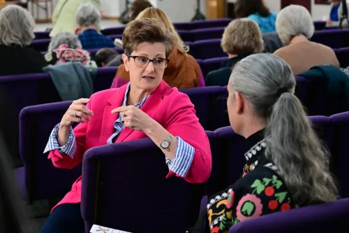 A woman with short brown hair and a bright pink jacket faces to the bottom right of the image to talk to another women with long gray hair who is facing her.