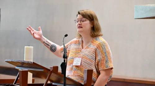 A woman stands at a pulpit with her arm outstretched and her eyes closed.