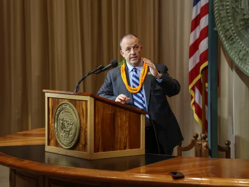 Man gesturing while standing at a lectern with a flag behind him