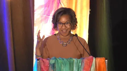 A woman speaks at a podium in front of a colorful backdrop.