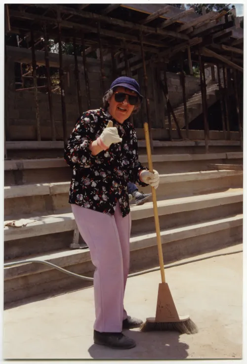 Margaret Purchase holds a broom in one hand and makes the "hang loose" sign with her other. She is wearing sunglasses and a hat on her head and light purple jeans.