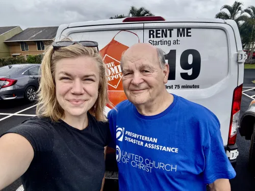 A smiling woman with sunglasses on her head shares a man wearing a blue t-shirt with Presbyterian Disaster Assistance on it.