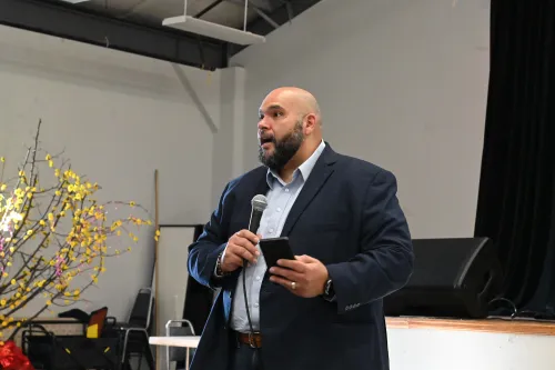 A man in a dark blazer speaks while holding a microphone.