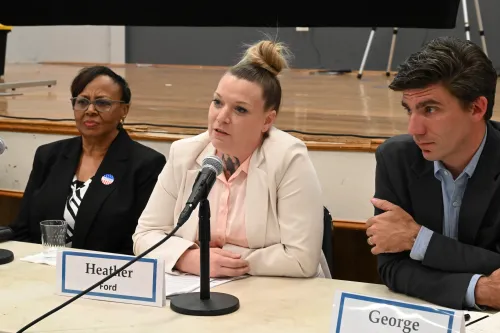 A woman in a beige jacket speaks at a microphone while part of a panel.