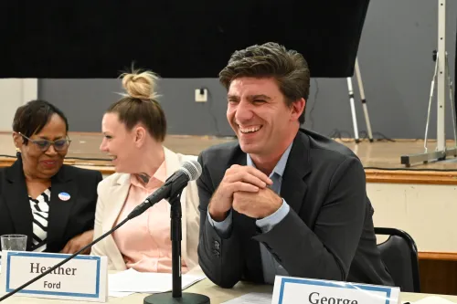 A manwith his hands clapsed smiles while seated with fellow panelists at a table.