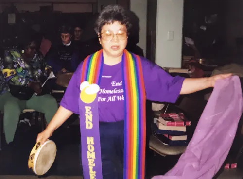 The Reverend Jean Kim wearing a purple "End Homelessness for all People" shirt, rainbow stole, holding tambourine while participating in worship.