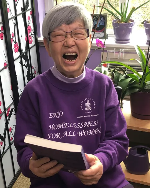The Rev. Jean Kim wearing her purple shirt, holding a book, and laughing aloud.