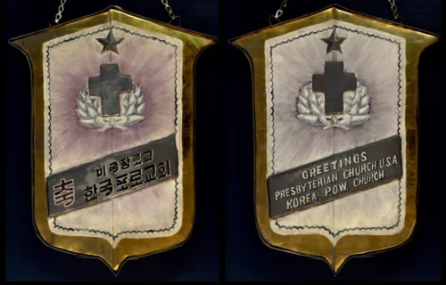 Plaque fashioned from beer cans. “Greetings Presbyterian Church U.S.A. Korea P.O.W. Church," The banner text sits below a thick black cross, which is topped by a five-pointed star and cushioned by a pillow of white feathers. 