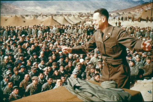 Harold Voelkel is at the right of the frame, his arm extended into the middle of the photograph, hand pointing a finger at the crowd on the left of the frame, who sit and listen to service.