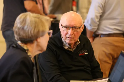 two seated people talk in foreground with other people standing behind them