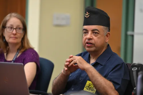 A portrait of a seated man wearing a hat, clasping his hands while speaking.