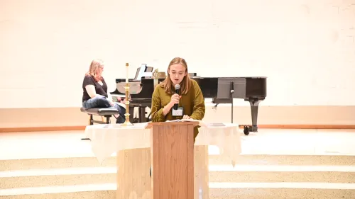 A speaker with blond hair stands at a podium; a pianist is seated in the background