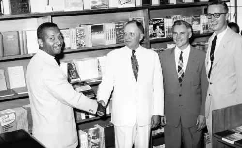 A black man at the left of the frame shakes hands with one of three white men to the right. All wear suits. Behind them are shelves full of books.