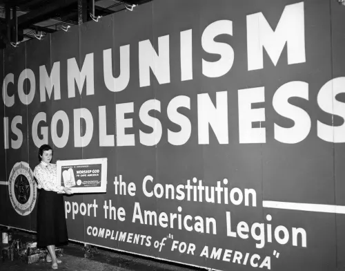 A large billboard that reads COMMUNISM IS GODLESSNESS. In front of the billboard stands a woman in a skirt, holing up a smaller illustration of the board.