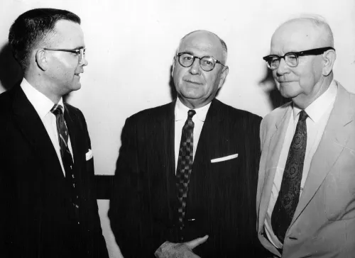 Three clergymen standing together in the center of the image, all three wearing suits and all three with glasses.