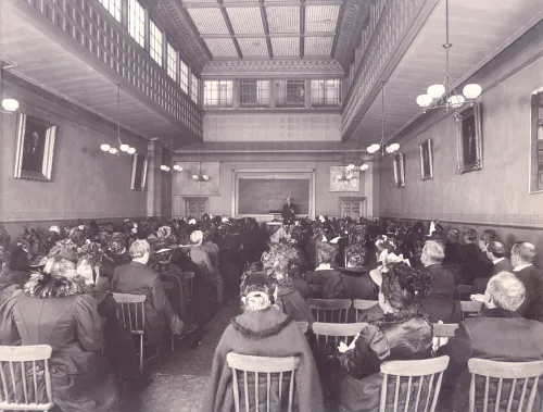 Photograph of a room filled with people sitting in chairs, at the front of the room is a man standing in front of a blackboard.
