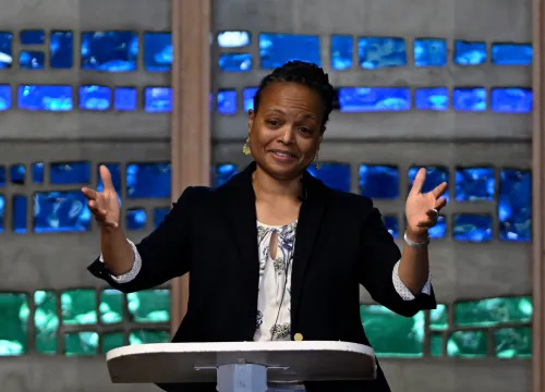A woman speaks at a lectern in front of blue and green stained glass.