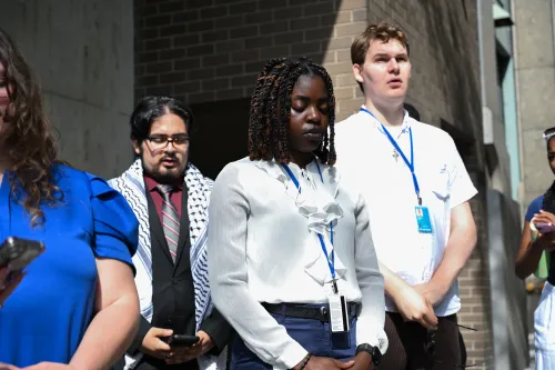 Three young adults bow their heads in prayer outside of a UN building. In the background, one young man wears a prayer shawl over his shoulders.