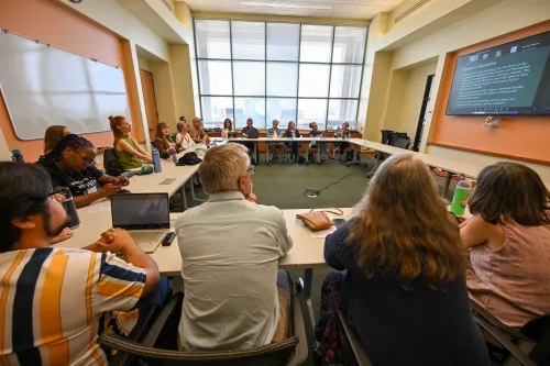 A group of people pack into a conference room with a large video screen and windows. 