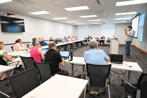 A person stands at a podium addressing a group seated at tables in a conference room, with some attendees using laptops.