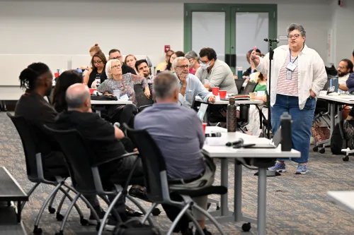 A woman stands in front of an audience at a microphone to ask a question of four people at a table.