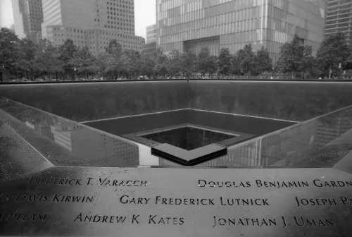 A black-and-white view of the 9/11 Memorial, showing engraved names on reflective pools surrounding a central void.
