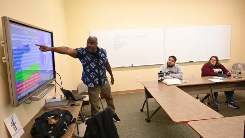 A man stands and points toward a screen on the lefthand side that talks about PCUSA policy on poverty. On the right, several young adults sitting at tables look at him attentively.
