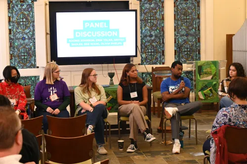 Six young adults sit in chairs side by side, with an audience in front of them. A man second from the right speaks into a mic while the others look on. Behind them, a screen says "panel discussion."