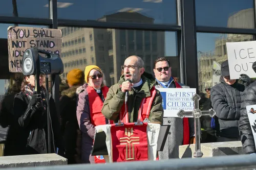 A man in a clergy collar speaks at a podium bearing the PC(USA) cross. Behind him stand a woman and man, also in clergy collars, and the man is holding a Presbyterian church sign. In the background, onlookers hold protest signs quoting scripturing and saying "ICE OUT."