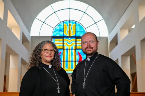 A man and a woman stand side by side in clergy collars and black shirts with silver cross necklaces. A stained glass window is behind them.