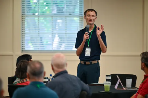 A person stands at the front of a room holding a microphone and gesturing while speaking to an audience seated at tables.