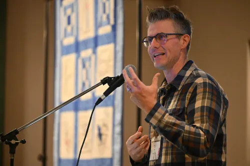 A person wearing glasses and a plaid shirt speaks into a microphone while gesturing with their hands.