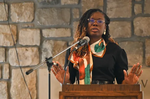 A person with long hair and glasses stands at a podium, speaking into a microphone while gesturing with open hands.
