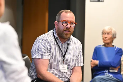 A person with glasses and a beard sits attentively in a meeting, wearing a striped shirt and a name badge, while another individual sits in the background.