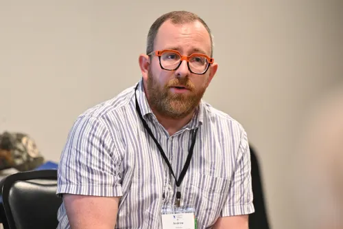 A person with a beard and orange-framed glasses sits in a chair, wearing a striped shirt and a lanyard with a name tag.