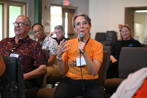 A person in an orange shirt speaks into a microphone while seated in a room with others listening.